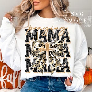 Puede incluir: Sudadera blanca con las palabras "praying MAMA" en letras negras y doradas. Un lazo con dise&ntilde;o de estampado de leopardo y una cruz dorada en el centro. Un art&iacute;culo con tem&aacute;tica de oto&ntilde;o.