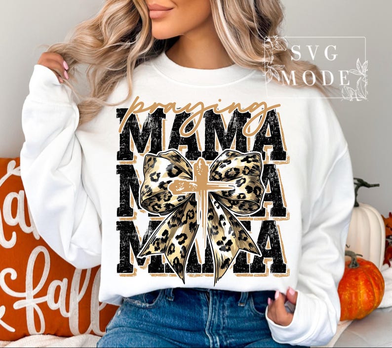 Puede incluir: Sudadera blanca con las palabras "praying MAMA" en letras negras y doradas. Un lazo con dise&ntilde;o de estampado de leopardo y una cruz dorada en el centro. Un art&iacute;culo con tem&aacute;tica de oto&ntilde;o.