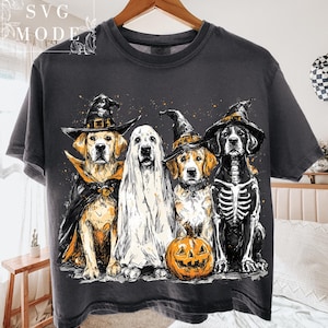 May include: Dark gray t-shirt featuring four dogs dressed in Halloween costumes. One dog is a ghost, another is a skeleton, and two wear witch hats and capes. A carved pumpkin sits in front of the dogs.