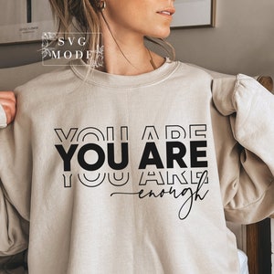 May include: A beige sweatshirt with the text "You Are You Are Enough" printed in black. The text is stacked vertically with the word "You" repeated three times.