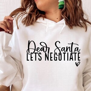 Pode incluir: Camisola de moletom branca com capuz com o texto "Caro Papai Noel, vamos negociar" em fonte de escrita cursiva preta com um pequeno s&iacute;mbolo de cora&ccedil;&atilde;o.