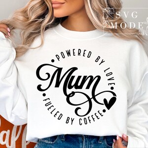 Może przedstawiać: Biała bluza z czarnym kołem z napisem "Powered by Love. Fueled by Coffee." z dużym, stylizowanym napisem "Mum".