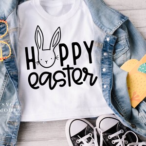 May include: White t-shirt with a black bunny and the text "Hoppy Easter". The shirt is laid out on a wooden surface with a denim jacket, a pair of black sneakers, and a yellow chick toy.