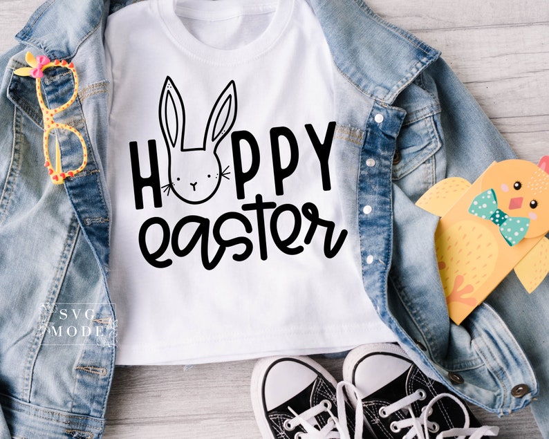May include: White t-shirt with a black bunny and the text "Hoppy Easter". The shirt is laid out on a wooden surface with a denim jacket, a pair of black sneakers, and a yellow chick toy.