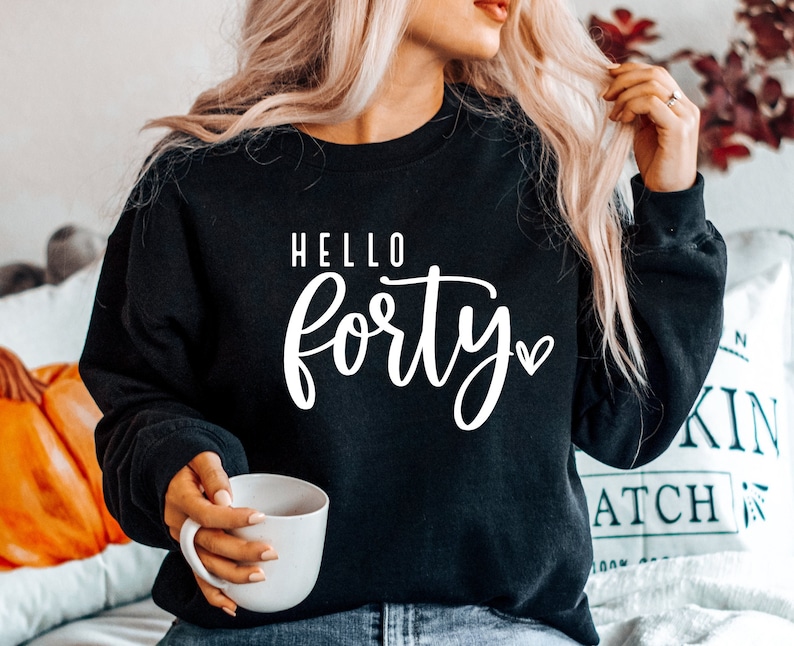 K&ouml;nnte beinhalten: Schwarzes Sweatshirt mit dem wei&szlig;en Schriftzug "HELLO forty". Ein kleines wei&szlig;es Herz befindet sich neben dem Wort "forty". Die Person, die das Sweatshirt tr&auml;gt, h&auml;lt eine wei&szlig;e Tasse.