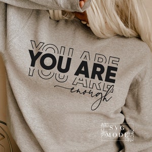 May include: A gray sweatshirt with the text "YOU ARE enough" printed in black. The text is stacked and stylized with overlapping letters.