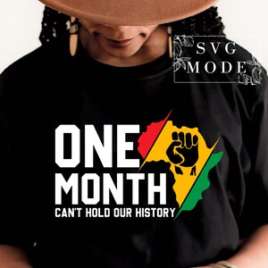May include: Black t-shirt with a white graphic design that says "One Month Can't Hold Our History". The graphic also includes a stylized image of a fist raised in the air, with red, yellow, and green colors.
