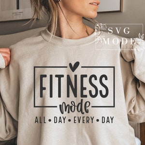 May include: A beige sweatshirt with the text "Fitness mode" in a black rectangle with a small heart above it. Below the rectangle is the text "All day every day".