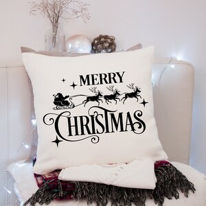 Puede incluir: Un coj&iacute;n navide&ntilde;o cuadrado de color crema con un gr&aacute;fico negro de Pap&aacute; Noel en un trineo tirado por renos, con las palabras "MERRY CHRISTMAS". El coj&iacute;n est&aacute; sobre una superficie blanca.
