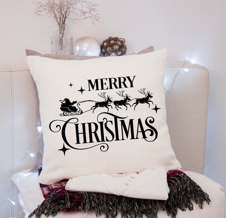 Puede incluir: Un coj&iacute;n navide&ntilde;o cuadrado de color crema con un gr&aacute;fico negro de Pap&aacute; Noel en un trineo tirado por renos, con las palabras "MERRY CHRISTMAS". El coj&iacute;n est&aacute; sobre una superficie blanca.
