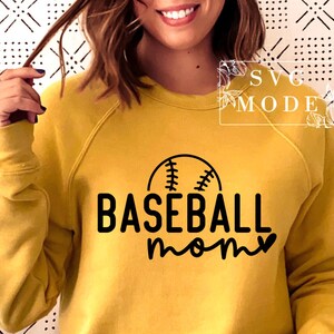 May include: Mustard yellow sweatshirt featuring a black baseball graphic and the words "BASEBALL mom". The design includes a baseball with stitching above the word "BASEBALL" and a heart replacing the "o" in "mom".
