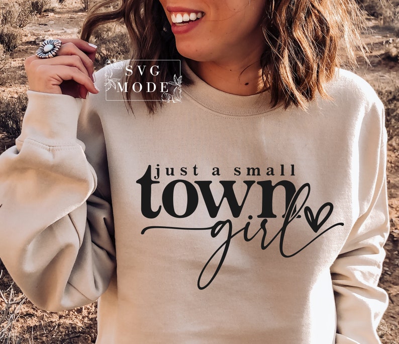 May include: A beige sweatshirt with the text "just a small town girl" printed on the front in black. A small heart is next to the text.