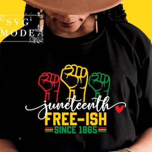 Könnte beinhalten: Schwarzes T-Shirt mit einem Grafikdesign zur Feier des Juneteenth. Das Design zeigt drei erhobene Fäuste in Rot, Gelb und Grün, mit den Worten "juneteenth FREE-ISH SINCE 1865" in Weiß und einem roten Herzen.