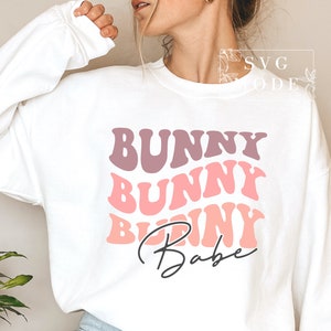 Może przedstawiać: Biała bluza z różowo-fioletowym nadrukiem, na którym widnieje napis "Bunny Bunny Bunny Babe" w retro czcionce.