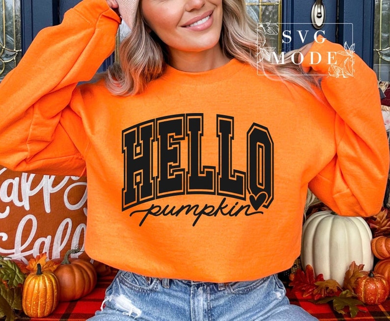 Może przedstawiać: Pomarańczowa bluza z czarnym napisem "HELLO pumpkin" z małym sercem po pumpkin. Bluzę nosi osoba.