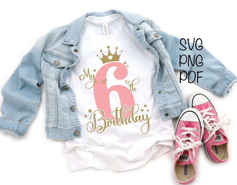 May include: A white t-shirt with a pink number 6 and gold lettering that says "My 6th Birthday" with a gold crown above the number. The shirt is on a blue denim jacket and pink sneakers.