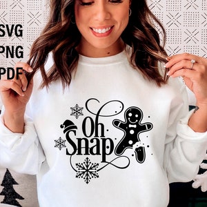 May include: White sweatshirt with black text that says "Oh Snap" and a gingerbread man graphic. The gingerbread man is wearing a red Santa hat.