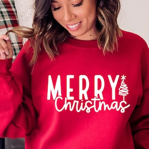 Puede incluir: Una sudadera roja con la inscripci&oacute;n blanca "MERRY Christmas" y un dise&ntilde;o estilizado de &aacute;rbol de Navidad. La sudadera es de color rojo liso y tiene cuello redondo.