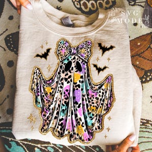 May include: A beige sweatshirt featuring a colorful ghost design with a leopard print bow, gold glitter accents, and black bats. The ghost has a colorful leopard print design with purple, yellow, and blue hues. The design is perfect for Halloween.