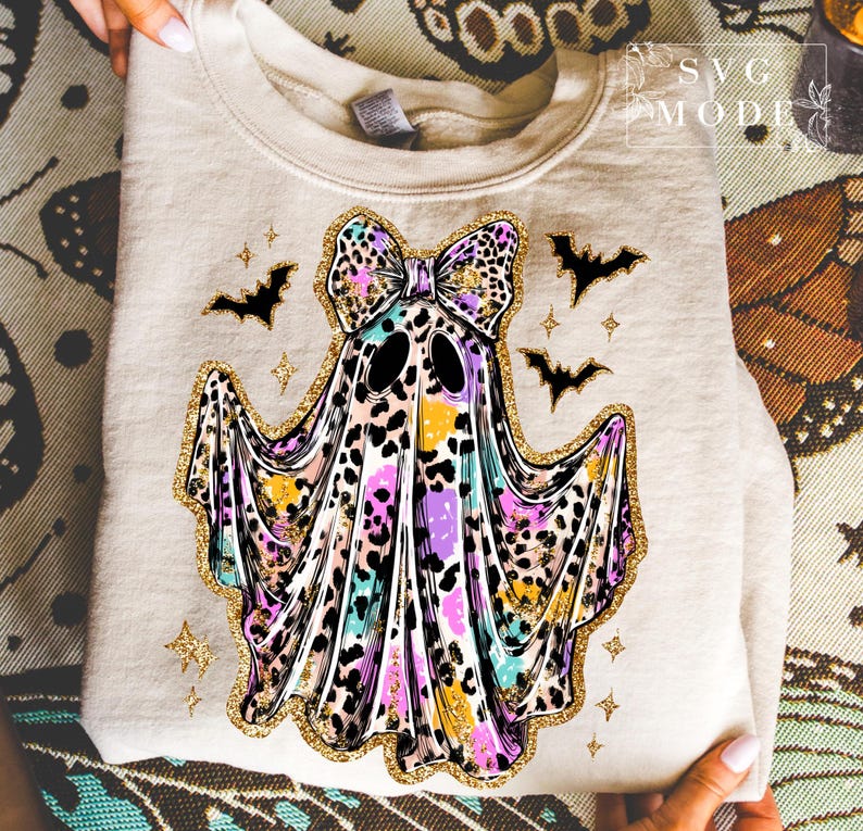 May include: A beige sweatshirt featuring a colorful ghost design with a leopard print bow, gold glitter accents, and black bats. The ghost has a colorful leopard print design with purple, yellow, and blue hues. The design is perfect for Halloween.