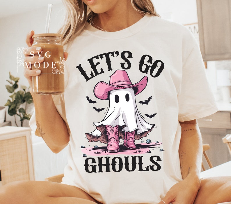 Może przedstawiać: Biała koszulka z nadrukiem ducha w r&oacute;żowym kowbojskim kapeluszu i butach. Na koszulce widnieje napis "Let's Go Ghouls".