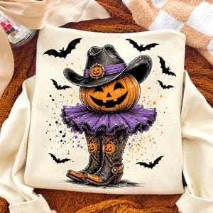 May include: A cream-colored sweatshirt featuring a graphic of a jack-o'-lantern wearing a black cowboy hat with a purple band, a purple ruffled skirt, and black cowboy boots with orange pumpkin accents, surrounded by black flying bats.