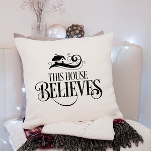 Puede incluir: Coj&iacute;n cuadrado color crema con la frase "THIS HOUSE BELIEVES" en escritura negra. El dise&ntilde;o incluye una peque&ntilde;a ilustraci&oacute;n de una casa. Parte de una acogedora decoraci&oacute;n del hogar.