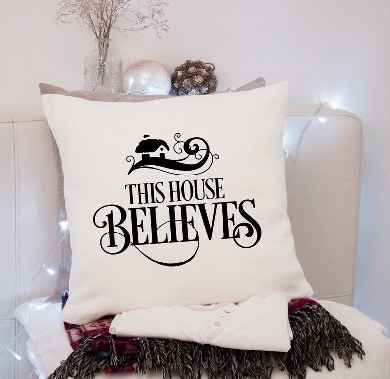 Puede incluir: Coj&iacute;n cuadrado color crema con la frase "THIS HOUSE BELIEVES" en escritura negra. El dise&ntilde;o incluye una peque&ntilde;a ilustraci&oacute;n de una casa. Parte de una acogedora decoraci&oacute;n del hogar.