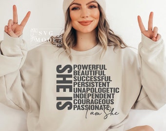 May include: A beige sweatshirt with the text "SHE IS" in black, followed by a list of words describing positive qualities, including "powerful", "beautiful", "successful", "persistent", "unapologetic", "independent", "courageous", and "passionate". The text "I am She" is written in cursive below the list.