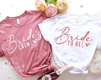May include: Two t-shirts, one dusty rose and one white, are displayed. The dusty rose shirt reads "Bride's Crew" in white script, and the white shirt says "Bride to Be" in red script. Both have a heart symbol. Flowers and a bottle of champagne are in the background.