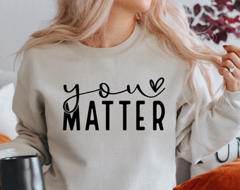 Könnte beinhalten: Ein hellgrauer Pullover mit dem Text "you matter" in schwarzen Buchstaben mit einem kleinen Herzen über dem Wort "you".