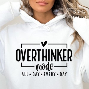 May include: White hoodie with the text "OVERTHINKER mode ALL DAY EVERY DAY" in black. The design includes a heart symbol above the word "OVERTHINKER". The hoodie has a drawstring hood and long sleeves.