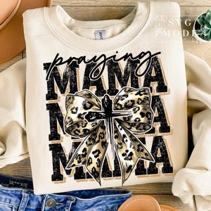 Puede incluir: Sudadera color crema con las palabras "Praying Mama" en negro y un dise&ntilde;o de lazo con estampado de leopardo y una cruz. La sudadera es de estilo casual y c&oacute;modo.