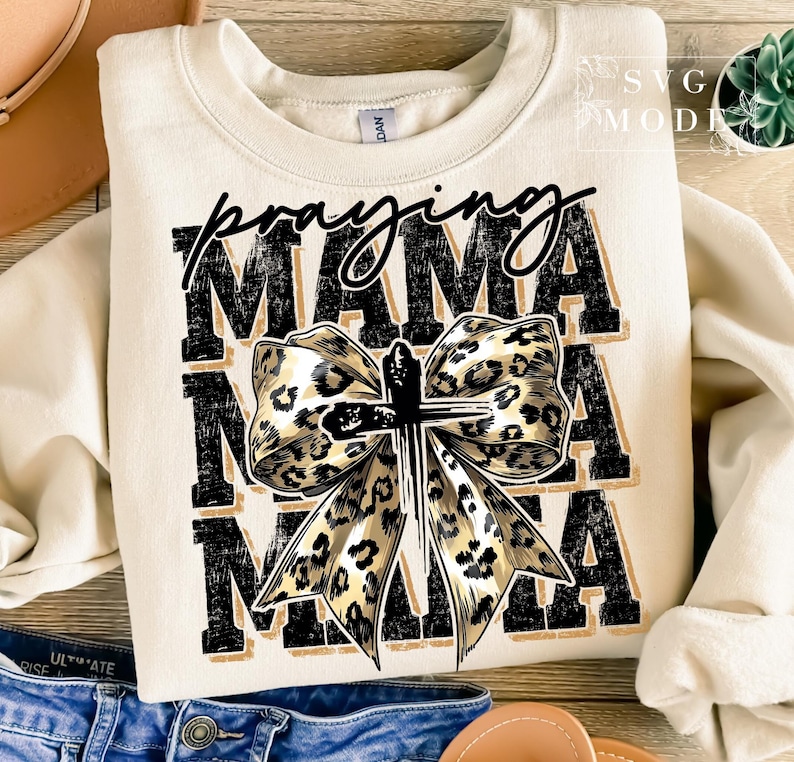 Puede incluir: Sudadera color crema con las palabras "Praying Mama" en negro y un dise&ntilde;o de lazo con estampado de leopardo y una cruz. La sudadera es de estilo casual y c&oacute;modo.