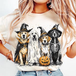 May include: A cream-colored t-shirt featuring a Halloween-themed graphic. The graphic depicts four dogs dressed in costumes: a witch, a ghost, a witch, and a skeleton, with a carved pumpkin. The design has a watercolor effect.