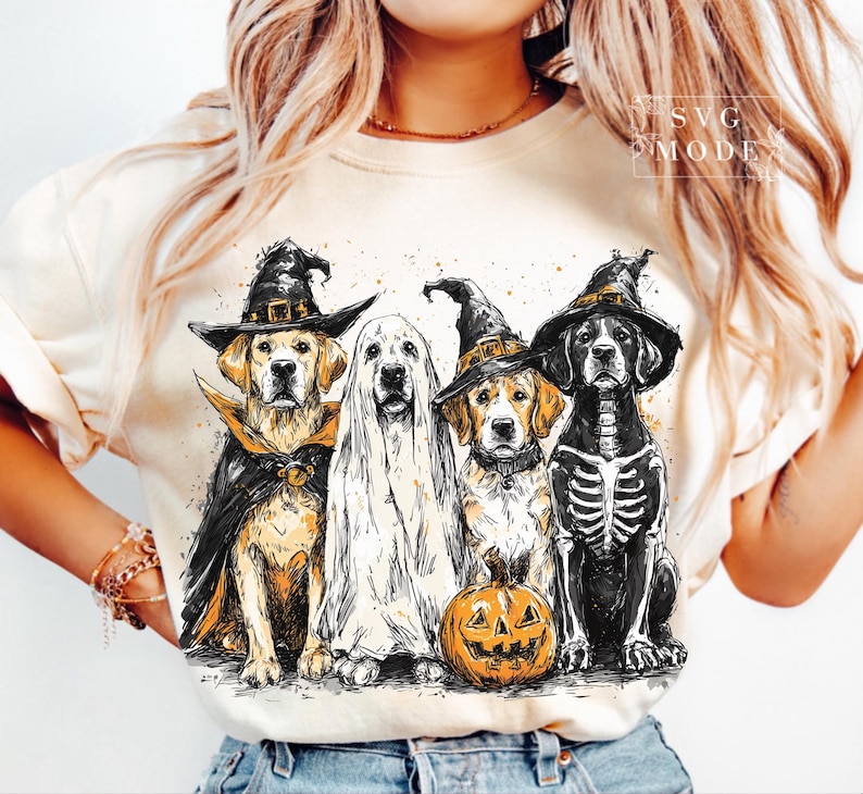 May include: A cream-colored t-shirt featuring a Halloween-themed graphic. The graphic depicts four dogs dressed in costumes: a witch, a ghost, a witch, and a skeleton, with a carved pumpkin. The design has a watercolor effect.