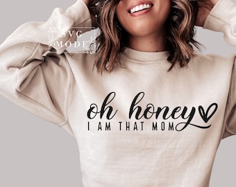 Puede incluir: Sudadera beige con la frase "oh honey I AM THAT MOM" en escritura negra. La palabra "honey" tiene un símbolo de corazón. La sudadera tiene cuello redondo y mangas largas.