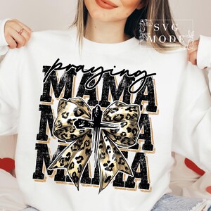 Puede incluir: Sudadera blanca con la inscripci&oacute;n "praying MAMA" en negro y dorado. La palabra "praying" est&aacute; en cursiva, y "MAMA" est&aacute; en negrita, en letras de bloque. Un lazo con estampado de leopardo con una cruz en el centro aparece debajo del texto.