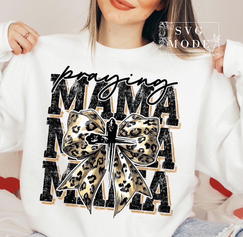 Puede incluir: Sudadera blanca con la inscripci&oacute;n "praying MAMA" en negro y dorado. La palabra "praying" est&aacute; en cursiva, y "MAMA" est&aacute; en negrita, en letras de bloque. Un lazo con estampado de leopardo con una cruz en el centro aparece debajo del texto.
