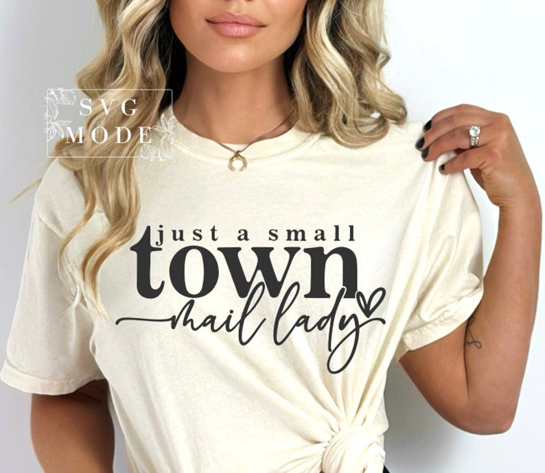 Peut inclure: Un t-shirt blanc avec le texte "Just a small town mail lady" en lettres noires avec un petit c&oelig;ur.