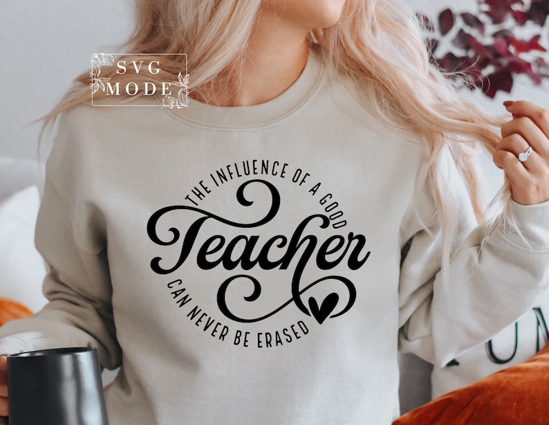May include: A white sweatshirt with black text that reads "The influence of a good teacher can never be erased". The text is in a cursive font and includes a small heart.