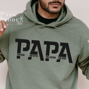 Peut inclure: Un sweat à capuche vert olive avec le mot "PAPA" en grandes lettres noires. En dessous, les phrases "THE MAN", "THE MYTH", "THE LEGEND" et "THE HERO" sont imprimées en noir.