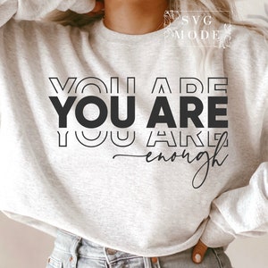 May include: A gray sweatshirt with the text "You are You are enough" printed in black. The text is in a bold, blocky font with a layered, outlined effect.