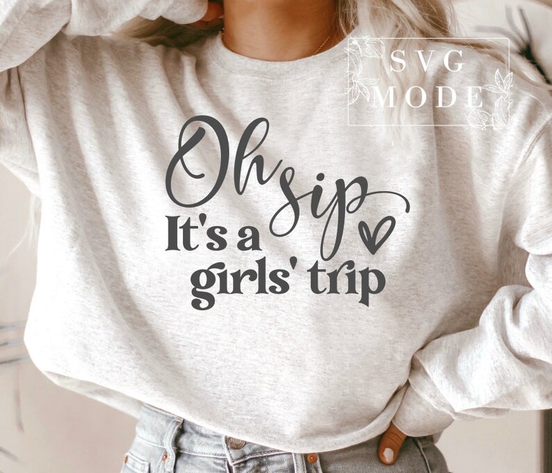K&ouml;nnte beinhalten: Ein grauer Pullover mit dem Text "Oh sip it's a girls' trip" in schwarzer Schrift mit einem kleinen Herz.
