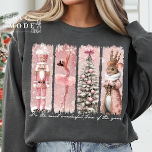 K&ouml;nnte beinhalten: Ein dunkelgrauer Pullover mit einem abgenutzten Design im Vintage-Stil, das einen rosafarbenen Weihnachtsbaum, eine Ballerina, einen Nussknacker und eine Maus mit Krone zeigt. Der Text "It's the most wonderful time of the year" ist unter dem Design gedruckt.