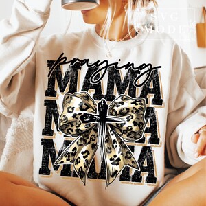 Puede incluir: Sudadera color crema con las palabras "Praying Mama" en escritura negra y letras de bloque. Un gran lazo con un dise&ntilde;o de estampado de leopardo y una cruz est&aacute; centrado en la parte delantera. La sudadera es de estilo casual y c&oacute;modo.