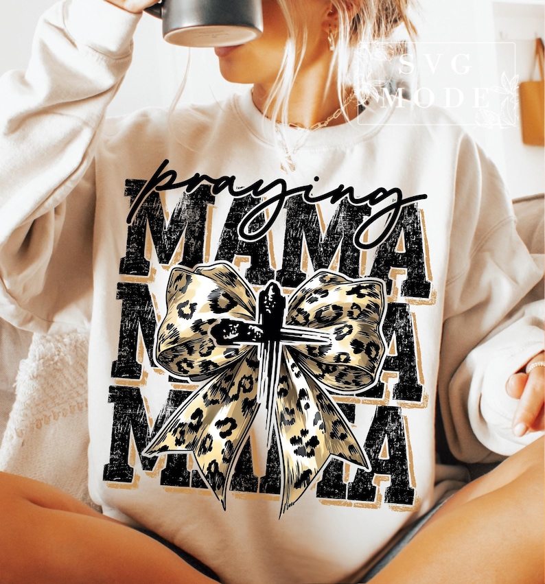 Puede incluir: Sudadera color crema con las palabras "Praying Mama" en escritura negra y letras de bloque. Un gran lazo con un dise&ntilde;o de estampado de leopardo y una cruz est&aacute; centrado en la parte delantera. La sudadera es de estilo casual y c&oacute;modo.
