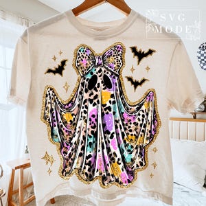 May include: Cream-colored t-shirt featuring a colorful ghost design with a leopard print bow and gold glitter accents. The ghost has black eyes and is surrounded by black bats and stars, perfect for Halloween.