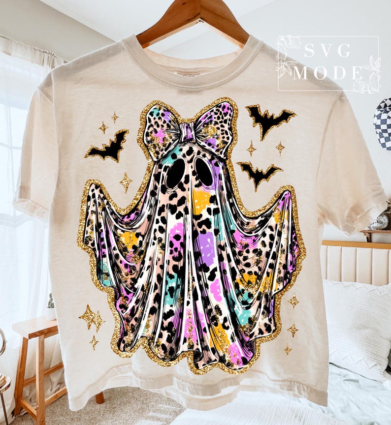 May include: Cream-colored t-shirt featuring a colorful ghost design with a leopard print bow and gold glitter accents. The ghost has black eyes and is surrounded by black bats and stars, perfect for Halloween.