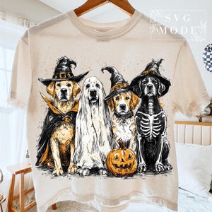 May include: Beige t-shirt featuring a Halloween-themed graphic of four dogs dressed in costumes. One dog is a vampire, another a ghost, a third a witch, and the last a skeleton. An orange pumpkin is in front of the witch dog.